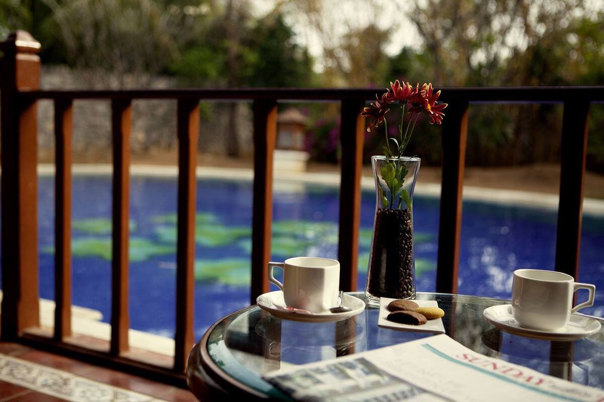 A glass table with two white cups, a vase with flowers, and a plate with cookies by a poolside.