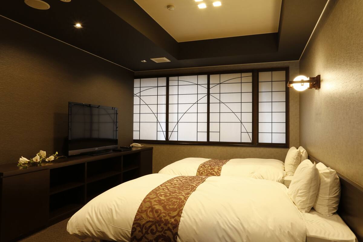 A hotel room with a large bed, a TV on a stand, a window with a view, and a wall-mounted light fixture.