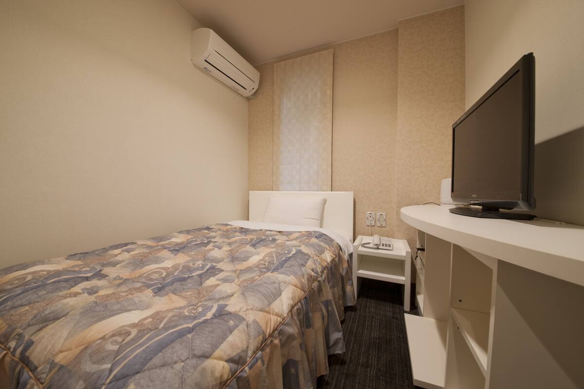A hotel room with a bed, a TV on a stand, a nightstand, and an air conditioning unit.