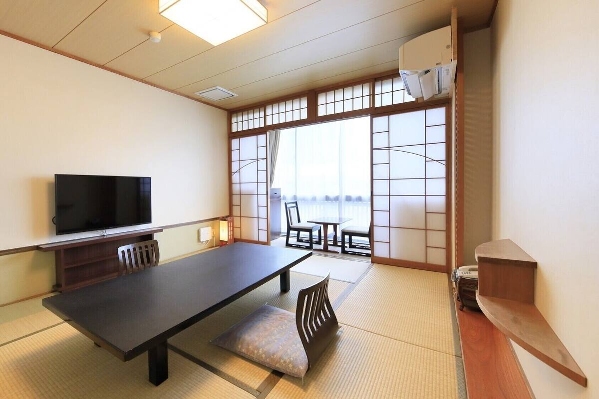A traditional Japanese room with tatami flooring, a television, a low table, and a sliding door leading to a balcony.