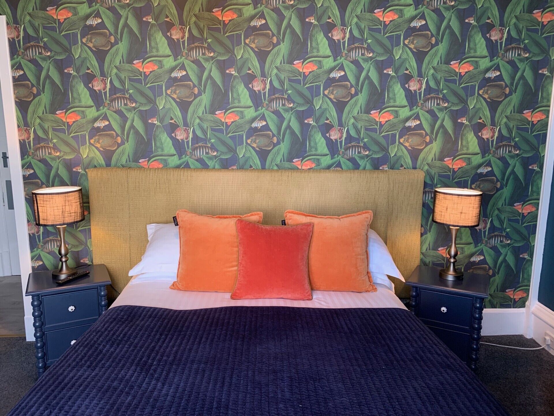 A bed with a dark blue quilt, two orange pillows, and a headboard with a light wood finish.