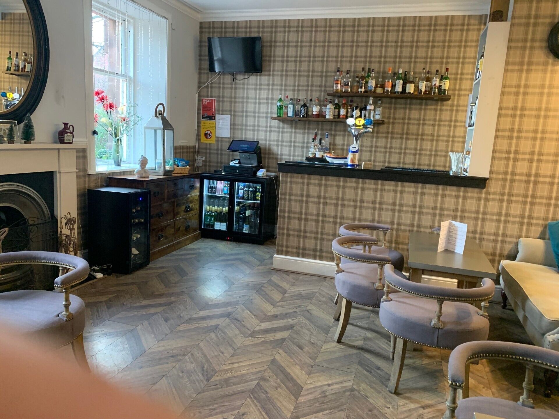 A bar area with a herringbone floor, a television mounted on the wall, and a variety of liquor bottles on shelves.