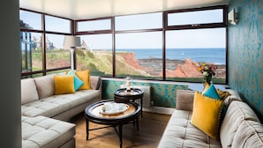 A room with a large window overlooking a coastal landscape, featuring a sofa, a small table with a tray of tea items, and a lamp. - Bayswell Park Hotel (Dunbar)