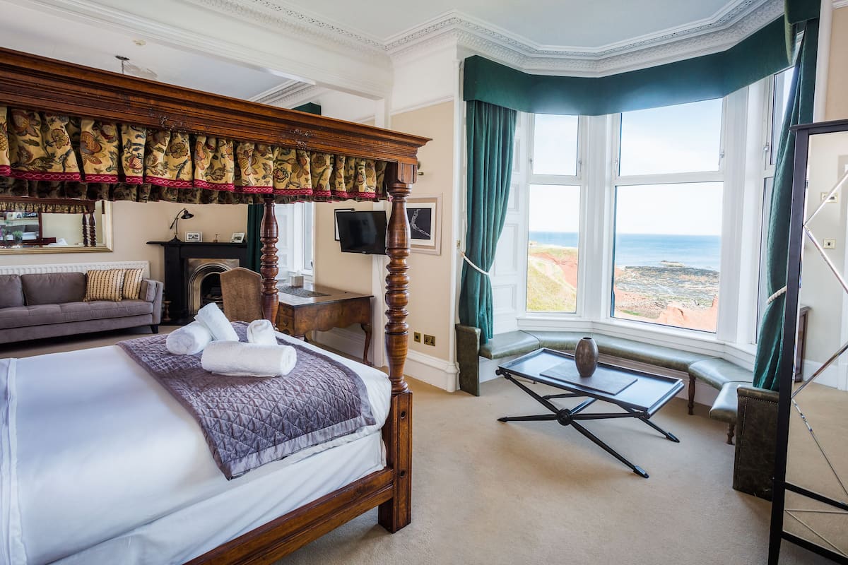 Uma cama com dossel com vista para o mar, um sofá, uma escrivaninha e uma mesinha perto da janela.