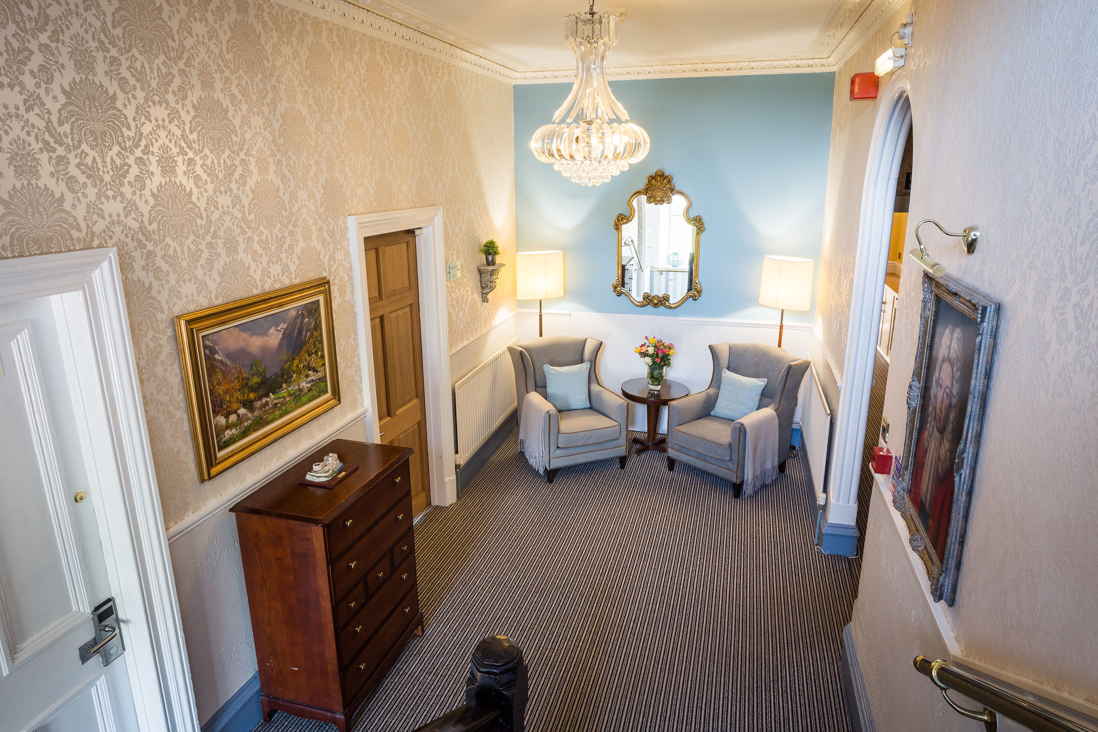 A hallway with a patterned carpet, a chandelier, a mirror, and a seating area with two armchairs and a small table.