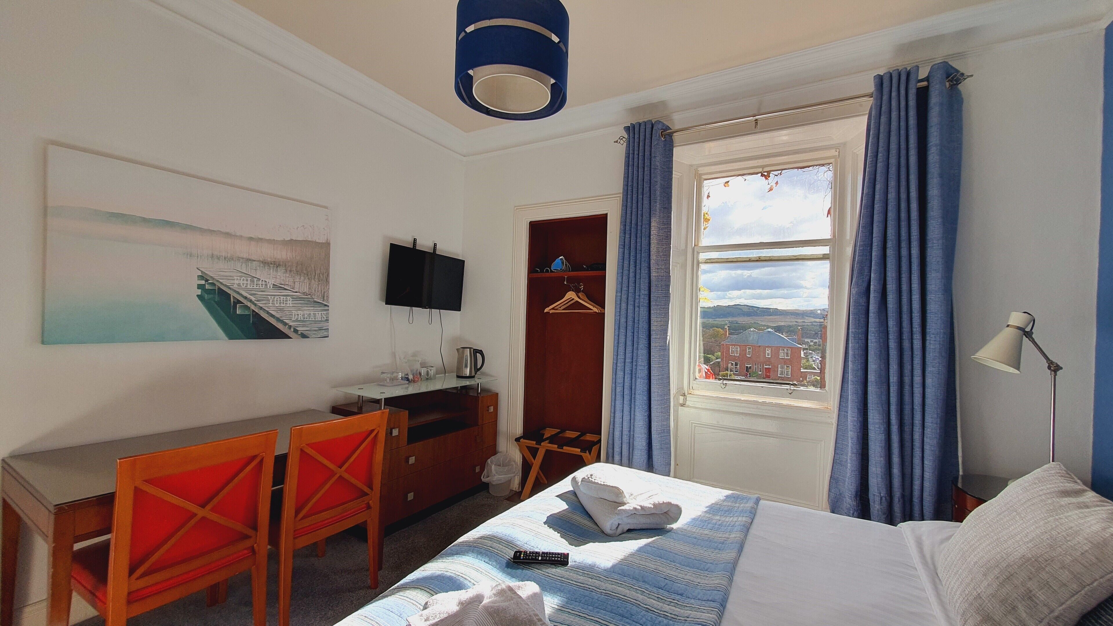 A hotel room with a bed, a desk with a chair, a television, a window with curtains, and a painting on the wall.
