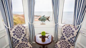Una habitación con dos sillas estampadas, una pequeña mesa con una planta y vista al mar a través de una ventana con cortinas.