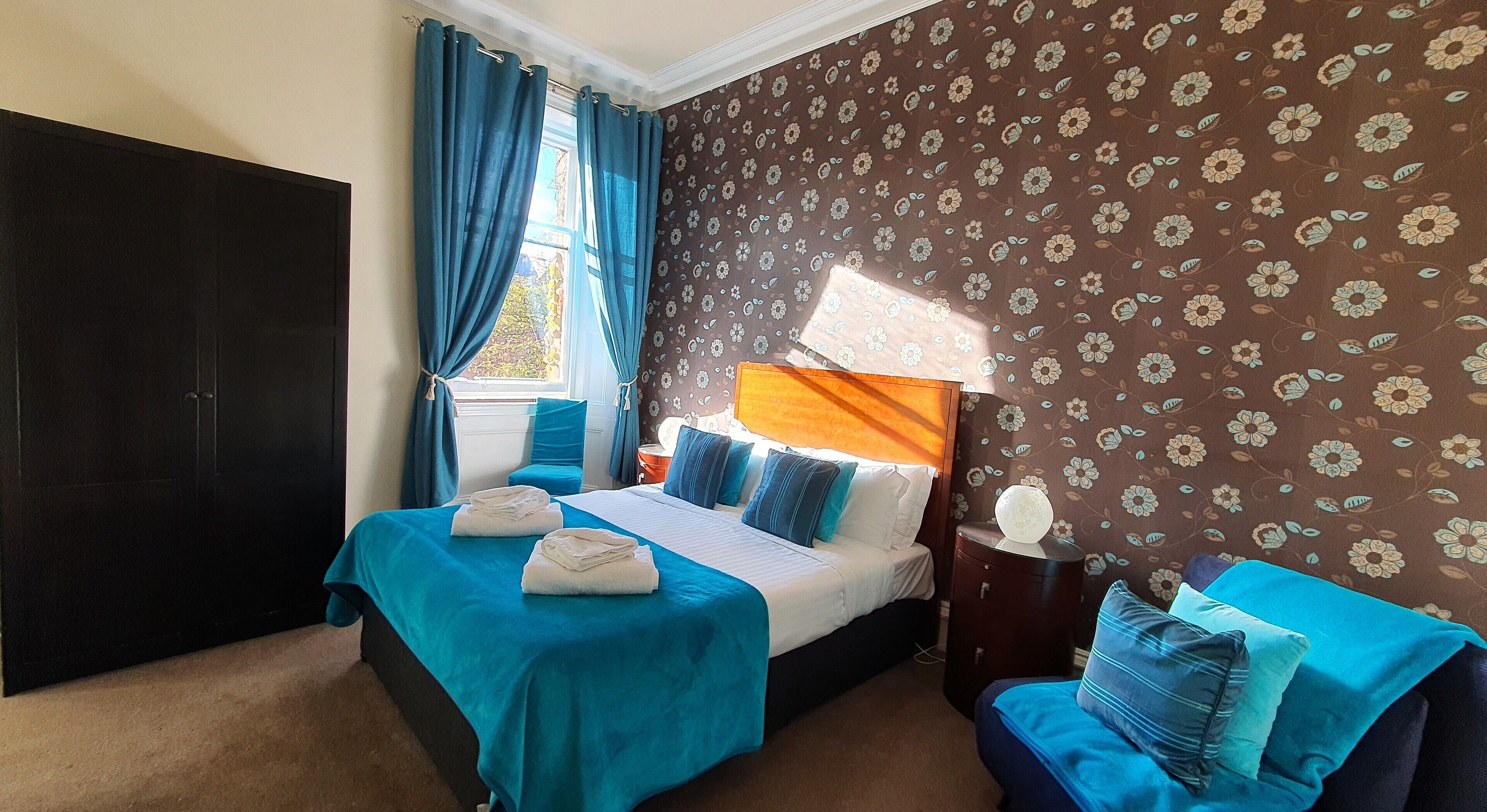 A hotel room with two beds, a large window with curtains, a floral wallpaper, and a nightstand with a lamp.