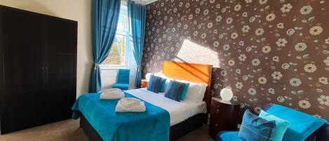 A hotel room with two beds, a large window with curtains, a floral wallpaper, and a nightstand with a lamp.