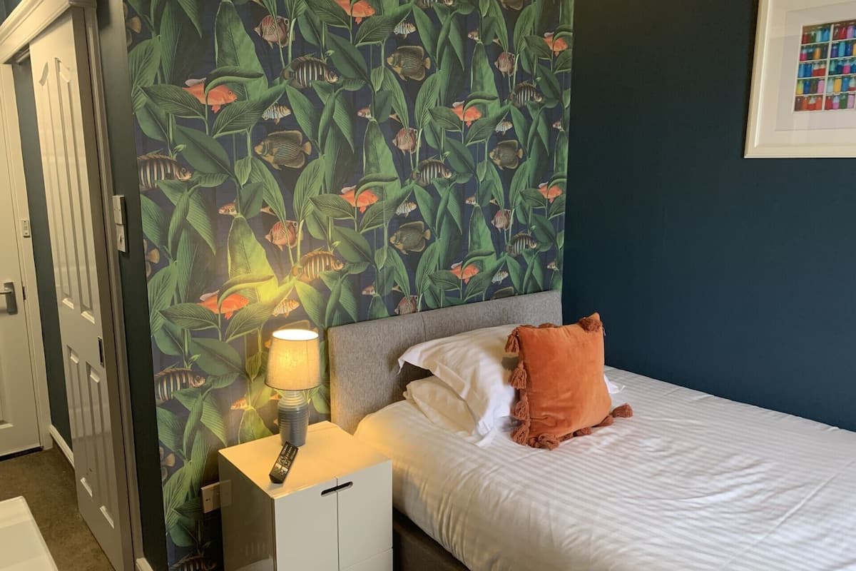 Quarto com cama, mesa de cabeceira, abajur e parede com padrão de peixes tropicais.