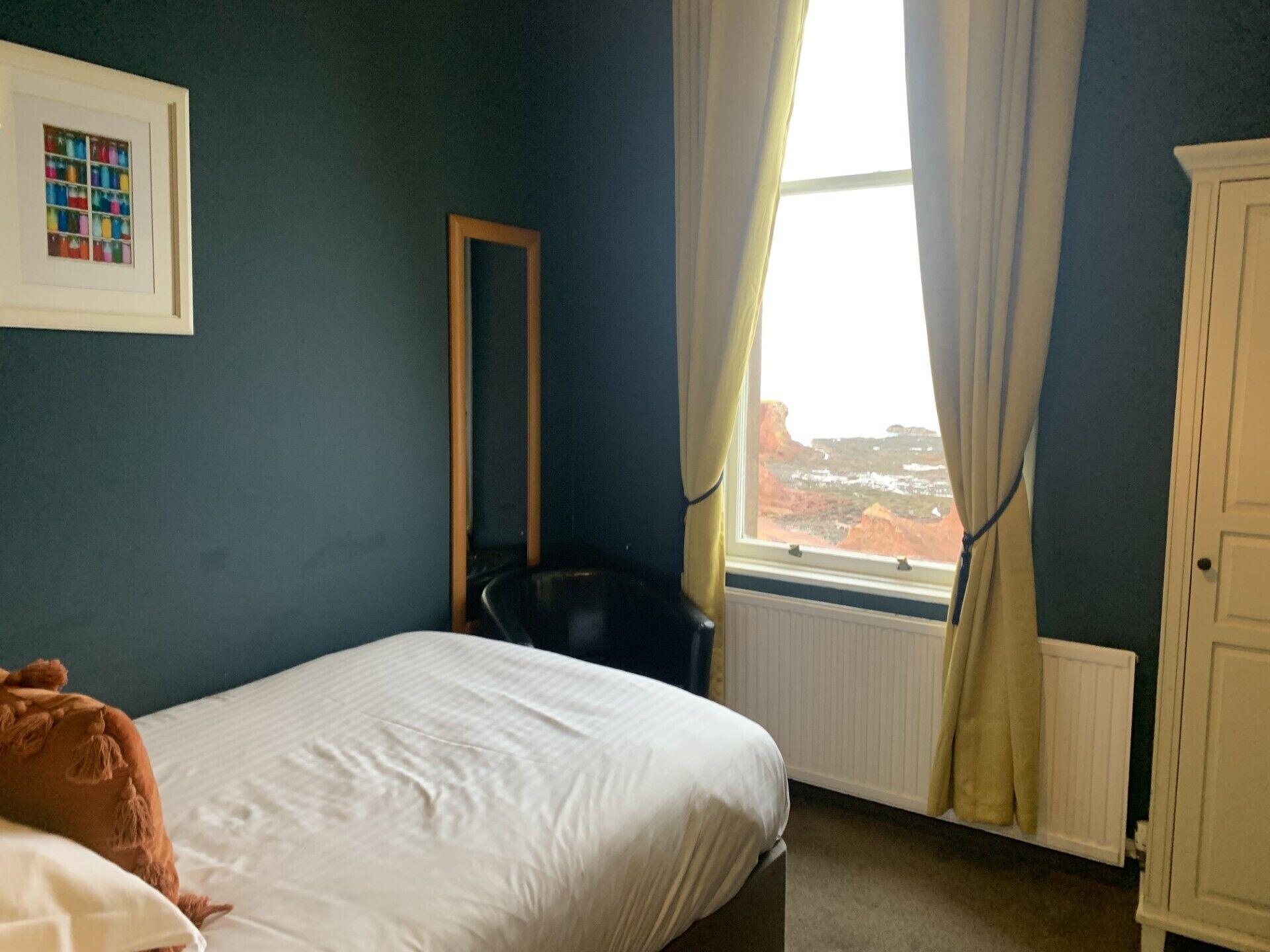 Ein Schlafzimmer mit Bett, Sessel, Heizkörper, Fenster mit Vorhängen und Schrank.