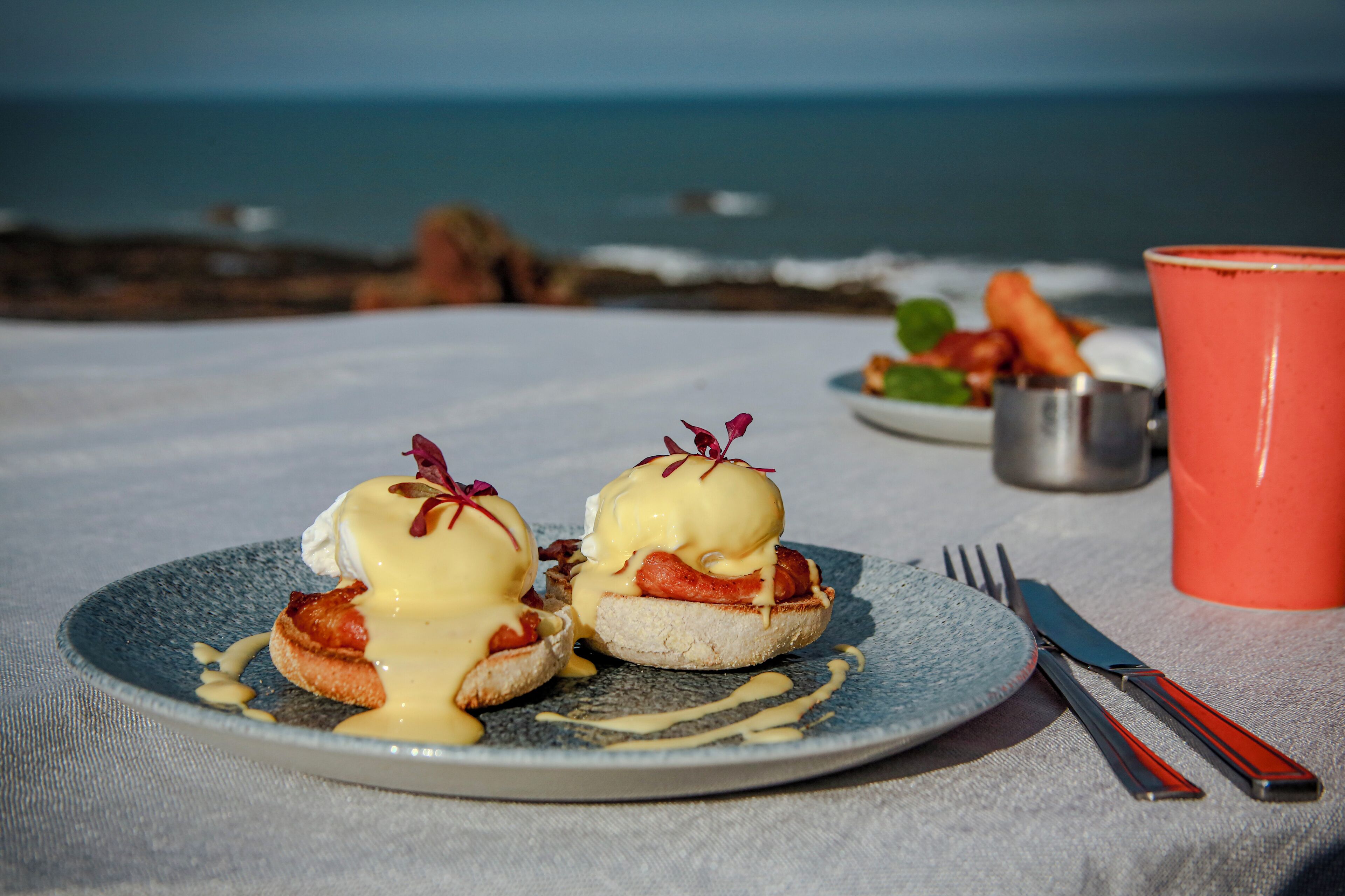 A table with a plate of eggs benedict, a red mug, and cutlery, set against a backdrop of the sea and rocky shore.