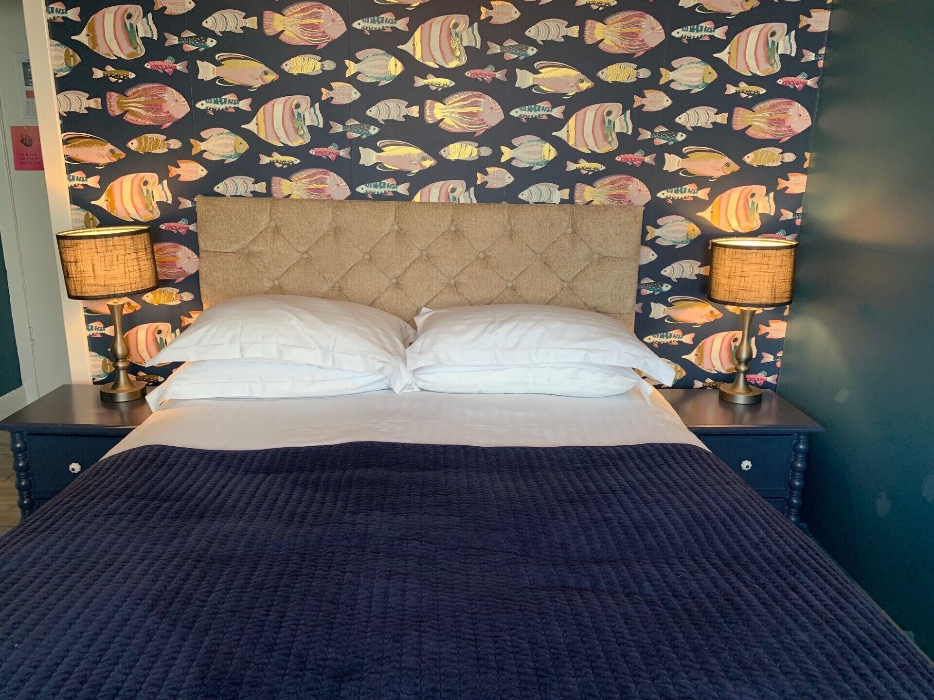 A bed with white pillows and a dark blue quilt, flanked by two nightstands with lamps, against a wall with a fish pattern wallpaper.