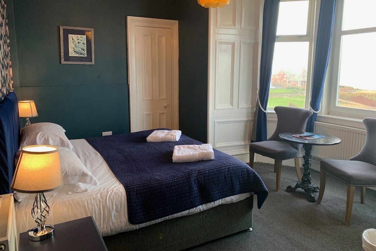 Quarto de hotel com cama, luminária de cabeceira, uma mesinha com cadeira, janela com cortinas e vista para um campo de golfe.
