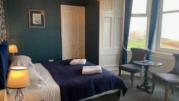 A hotel room with a bed, bedside lamp, a small table with a chair, a window with curtains, and a view of a golf course.