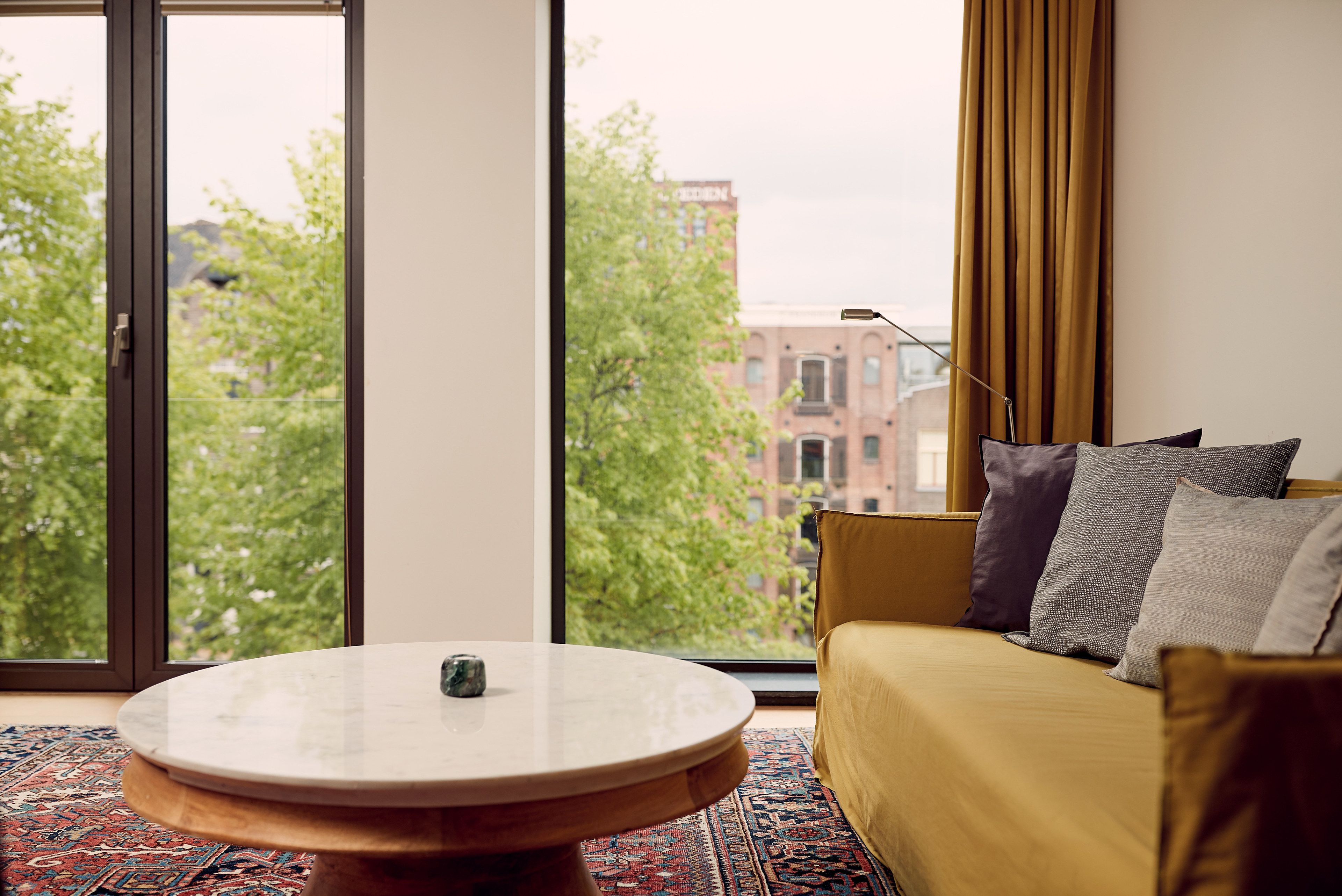 Una habitación con una mesa redonda, un sofá con cojines, una alfombra estampada y vistas a árboles y edificios a través de amplios ventanales.