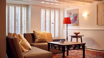 A living room with a sofa, a coffee table, and a lamp. There is a window with blinds and a painting on the wall.