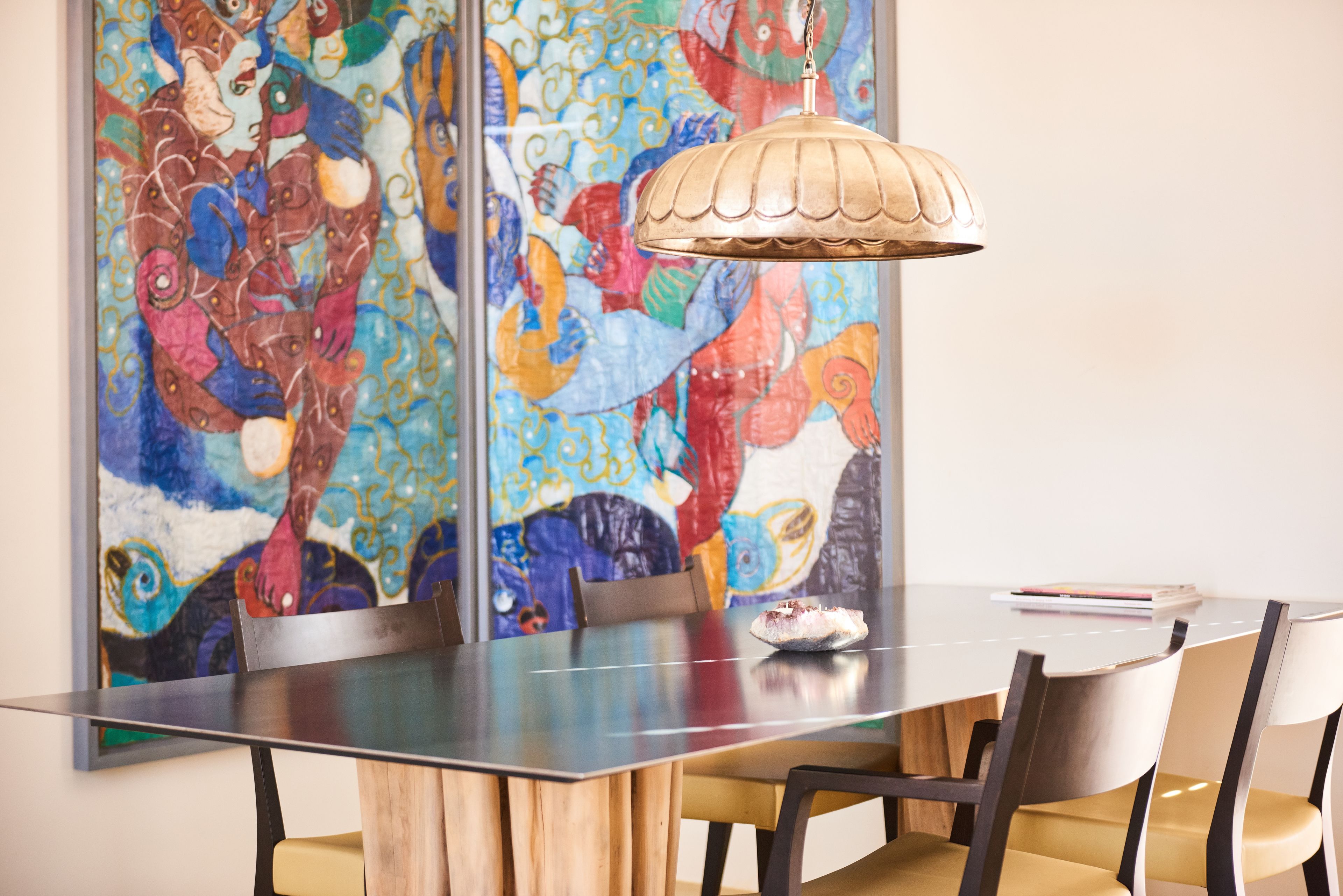 Un comedor con una mesa de madera, cuatro sillas, un cuadro abstracto y colorido en la pared, y una lámpara colgante.
