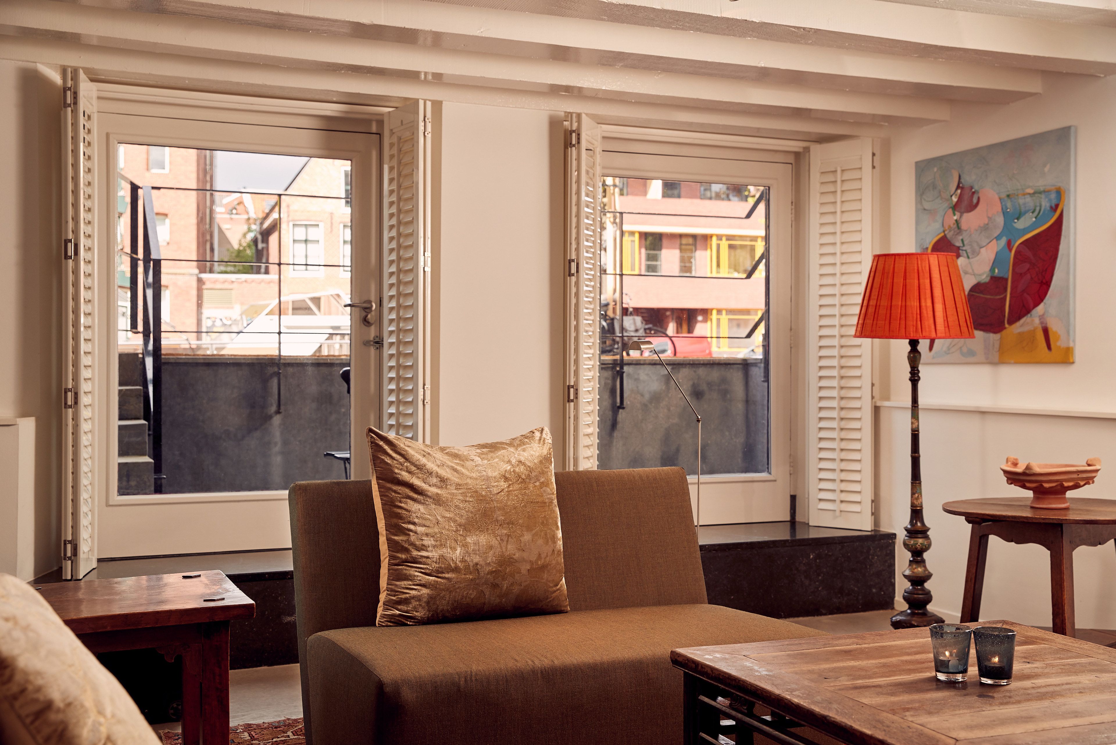 Un salon avec un canapé, une table et une lampe. Il y a deux fenêtres avec des volets et un tableau accroché au mur.