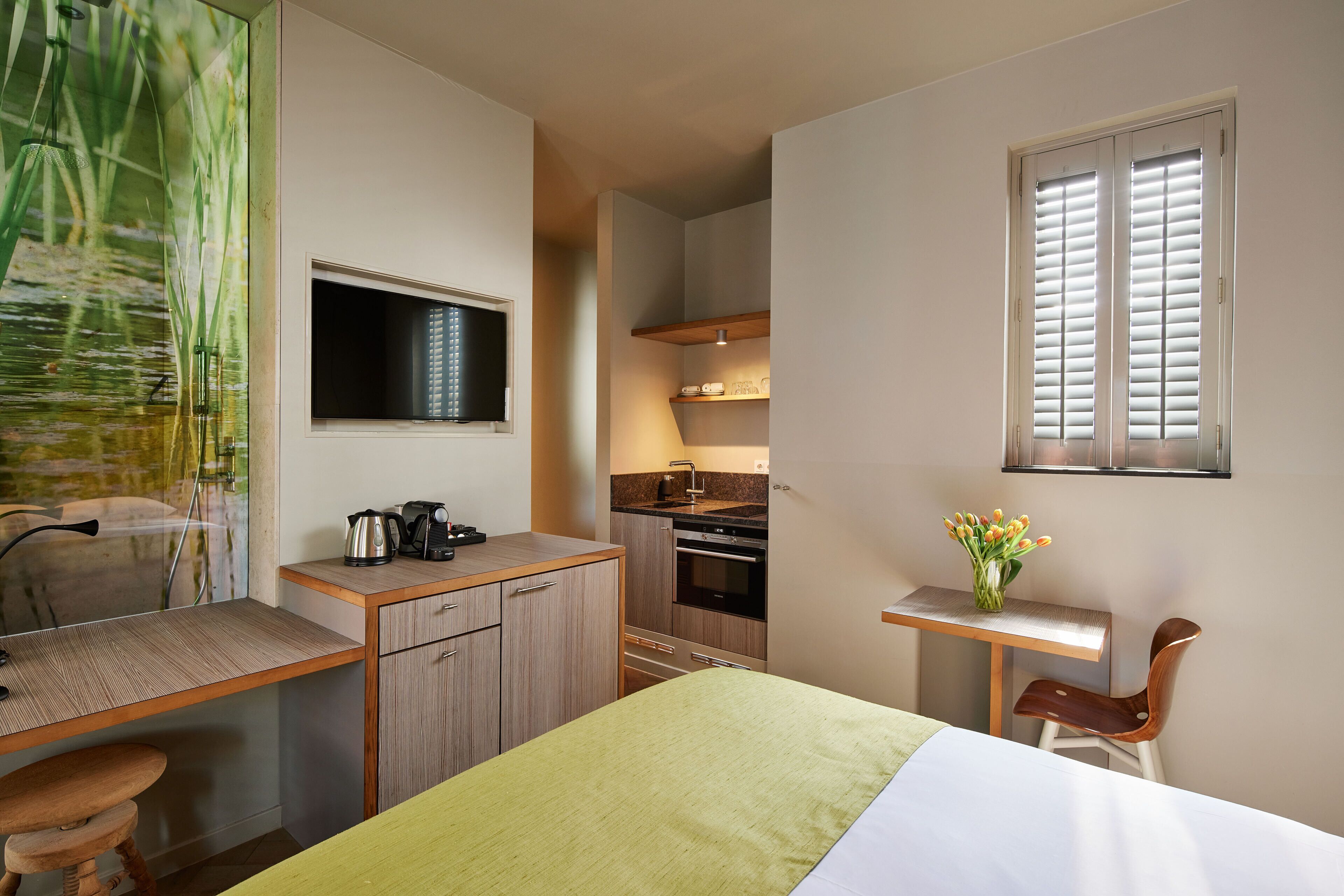 A compact hotel room with a bed, a desk, a chair, a small kitchenette, and a window with blinds.