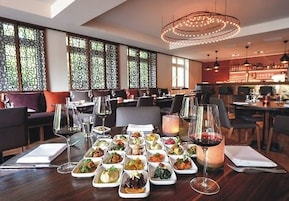 Un restaurante con varias mesas dispuestas, cada una con distintos platos y copas de vino, amplias ventanas con celosías decorativas y una moderna lámpara de araña.