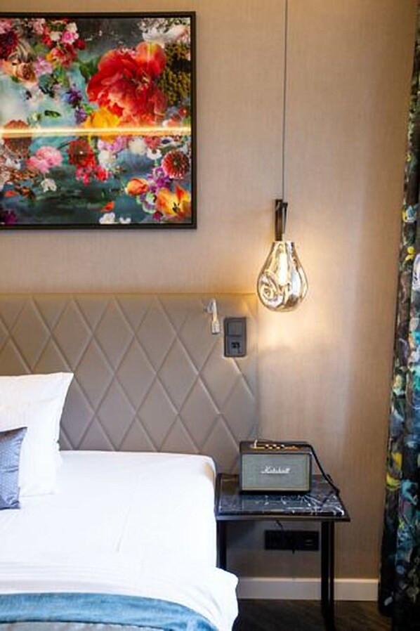 A hotel room with a bed, a wall-mounted artwork of flowers, a table with a radio, and a unique light fixture.