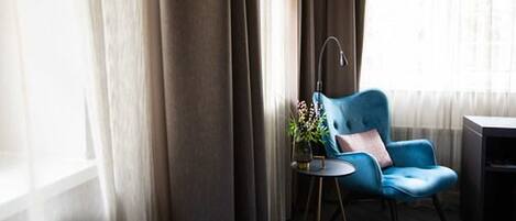 Una habitación con una silla azul, una mesita redonda pequeña con una lámpara y una ventana con cortinas.