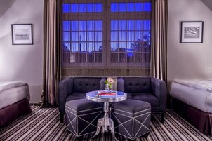 Una habitación de hotel con un ventanal grande, un sofá, una mesita redonda y dos camas.