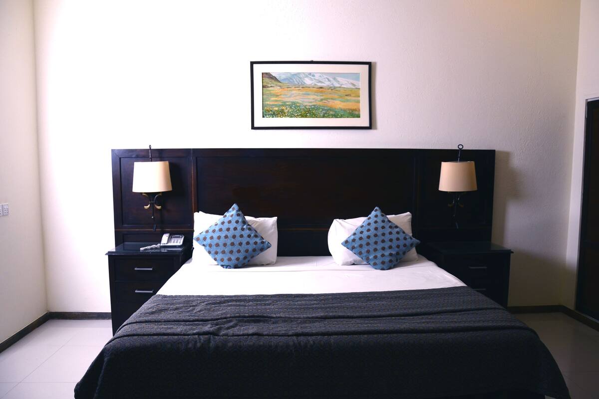 A hotel room with a bed, two nightstands, a telephone, and a framed landscape painting on the wall.