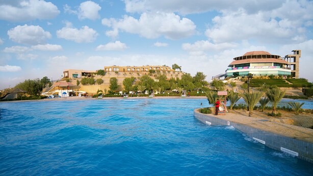 A large swimming pool with a view of a hillside building and a modern structure with balconies. - Dreamworld Resort, Hotel & Golf Course (Karachi)