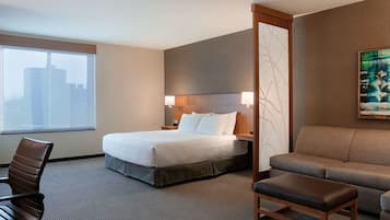 A hotel room with a bed, a sofa, a desk with a chair, a TV, and a painting on the wall.