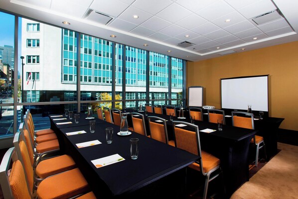 A conference room with a long table, chairs, a projector screen, and large windows offering a city view.