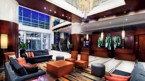 A spacious lobby with a modern design, featuring a central seating area with sofas and armchairs, a patterned carpet, and a staircase leading to an upper level.