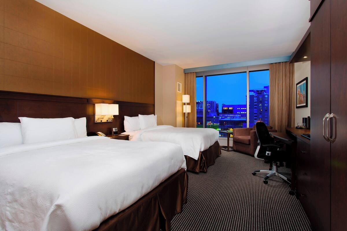 Habitación de hotel con dos camas, un escritorio, una silla y vista a la ciudad al anochecer.