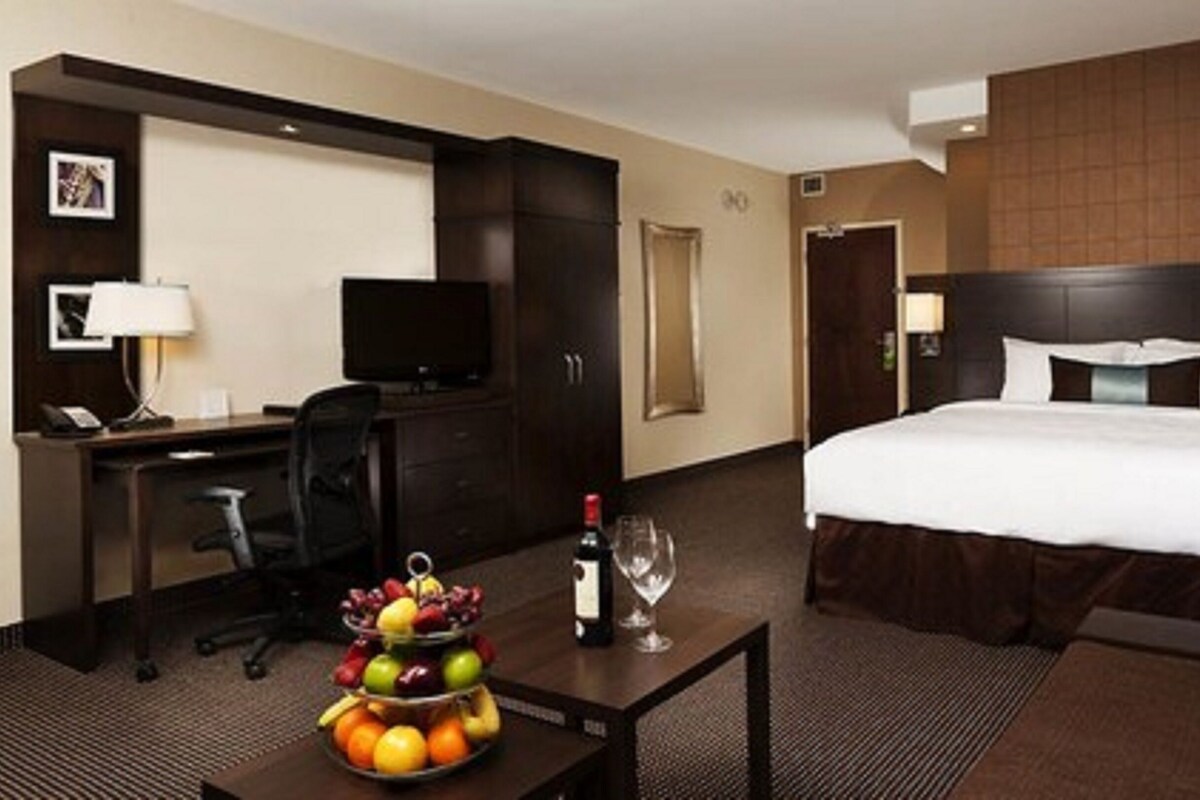 Une chambre d’hôtel avec un lit, un bureau avec un ordinateur, une chaise et un panier de fruits.