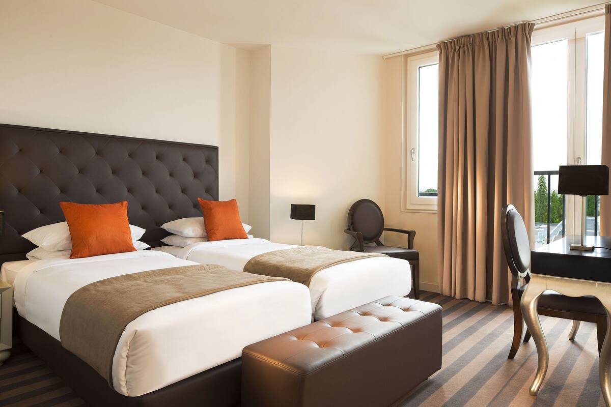 Ein Hotelzimmer mit zwei Betten, einem großen, gepolsterten Kopfteil, einem Hocker, einem Schreibtisch mit Stuhl und Blick ins Freie durch ein mit Vorhängen versehenes Fenster.