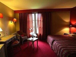 Une chambre d’hôtel avec un lit, un bureau, une chaise, une table avec un vase et une fenêtre avec des rideaux.