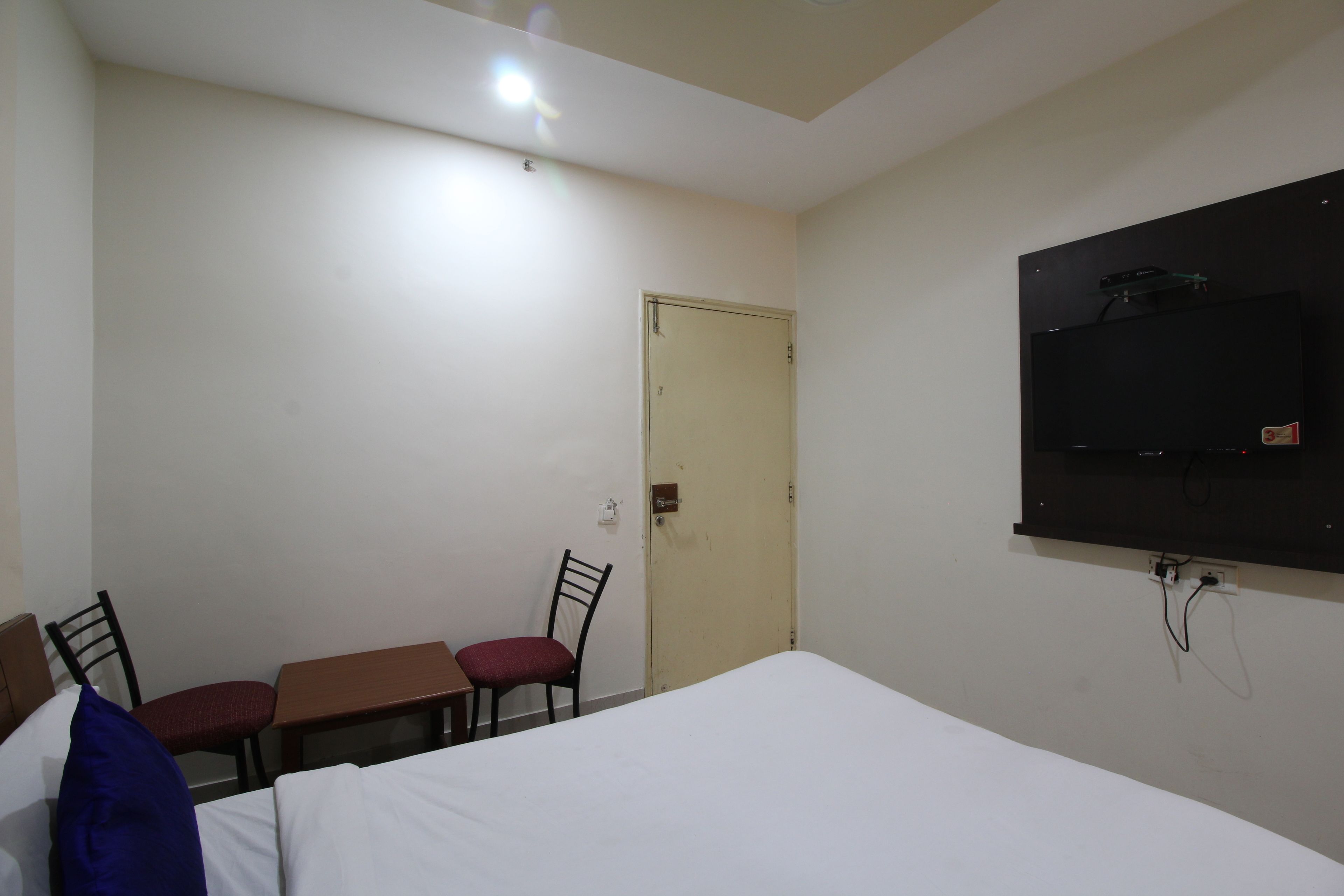 A hotel room with a bed, a desk, two chairs, a television mounted on the wall, and a door.