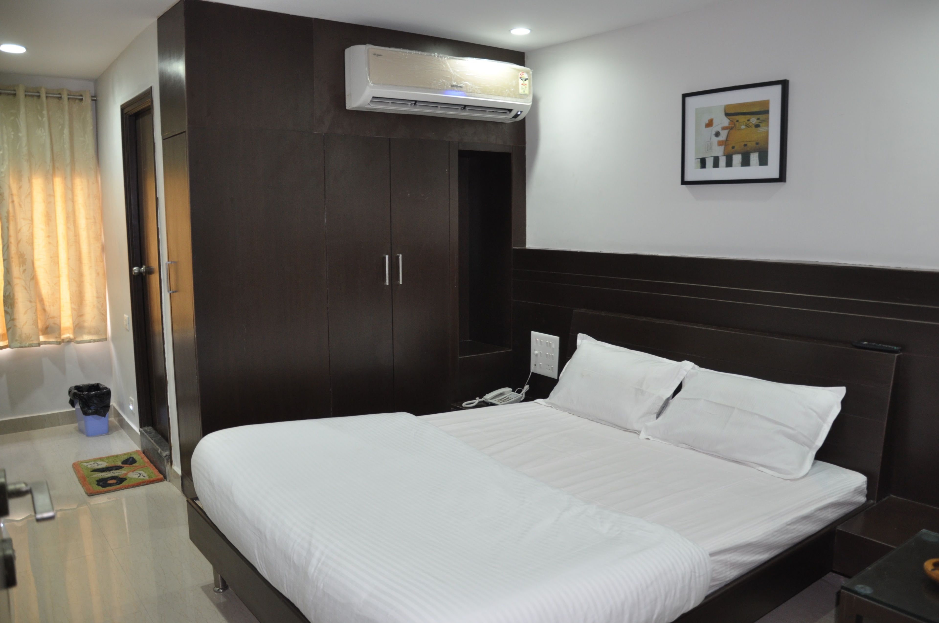 A hotel room with a large bed, a wardrobe, an air conditioner, a framed picture on the wall, and a trash bin.
