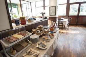 Ein reichhaltiges Buffet mit verschiedenen Speisen und Besteck, ein Essbereich mit Tischen sowie ein Blick ins Freie durch große Fenster.