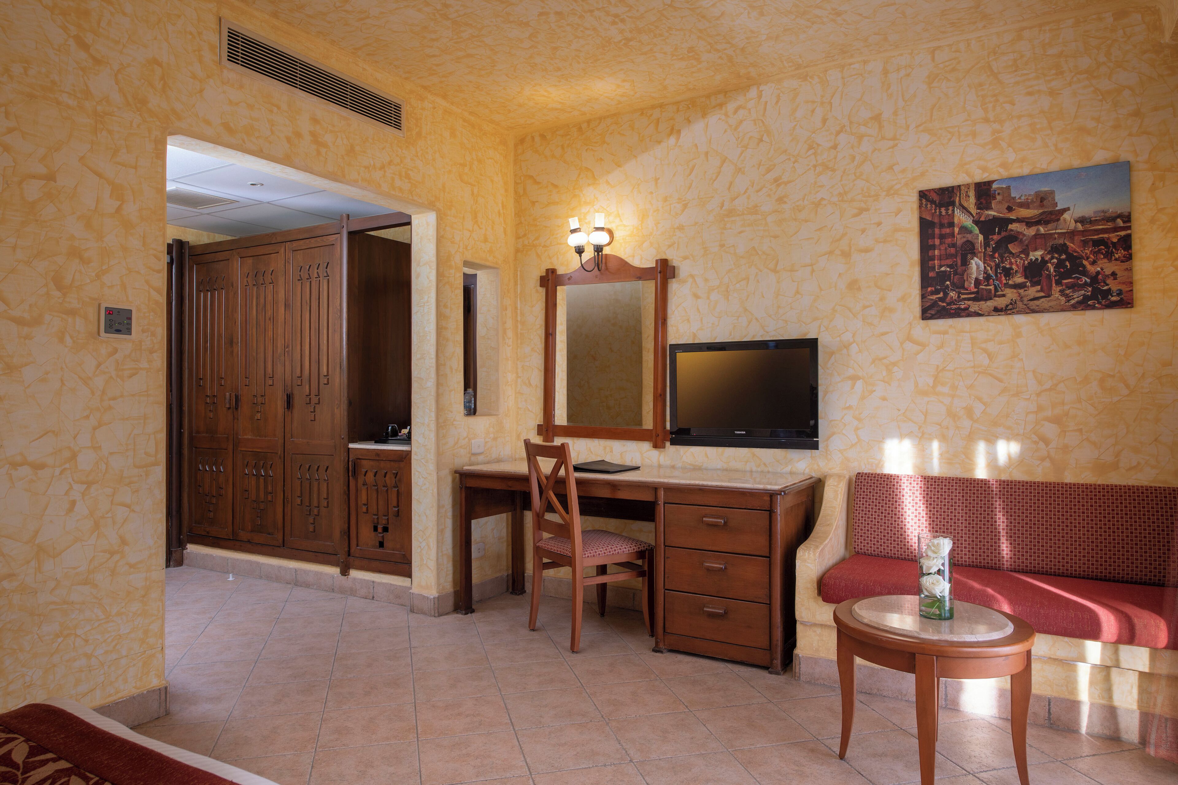 Una habitación con un escritorio de madera, una silla, un televisor, un sofá y una pequeña mesa con un jarrón.
