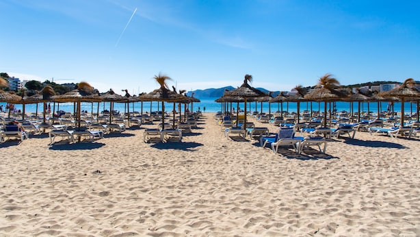 A beach with rows of sun loungers and thatched umbrellas, a clear blue sky, and a calm sea.