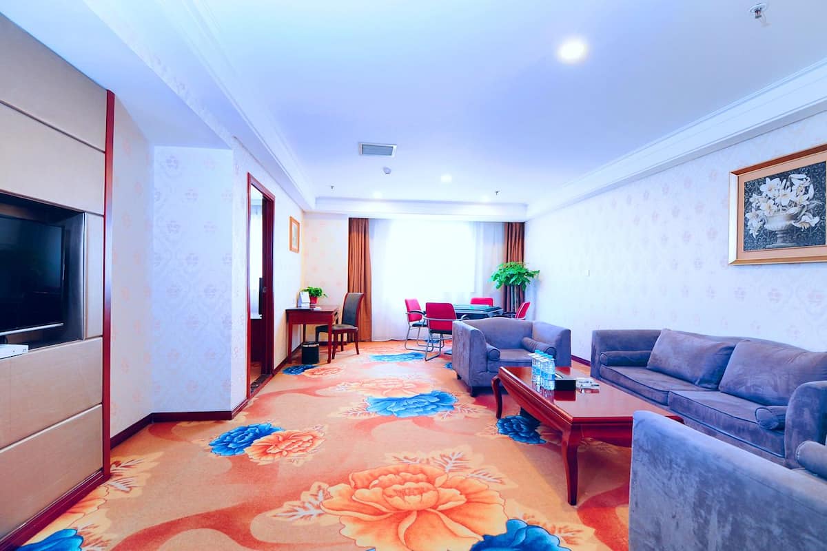 A hotel room with a TV, a sofa, a coffee table, a dining area with chairs, and a carpet with floral patterns.