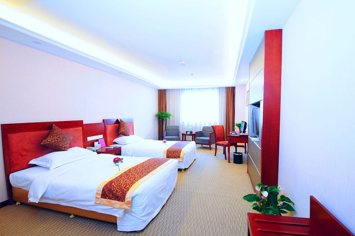 A hotel room with two beds, a desk, a chair, a TV, and a plant.