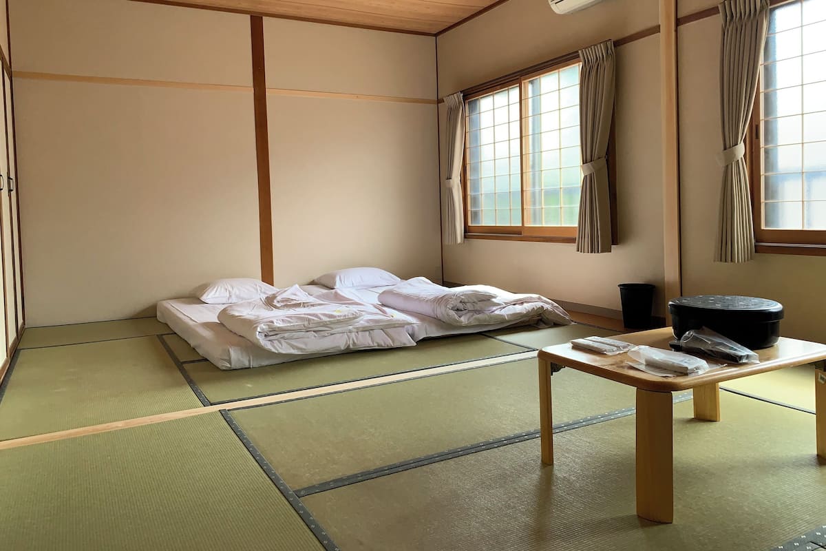 A traditional Japanese room with tatami flooring, a futon bed, a wooden table, and a window with curtains.