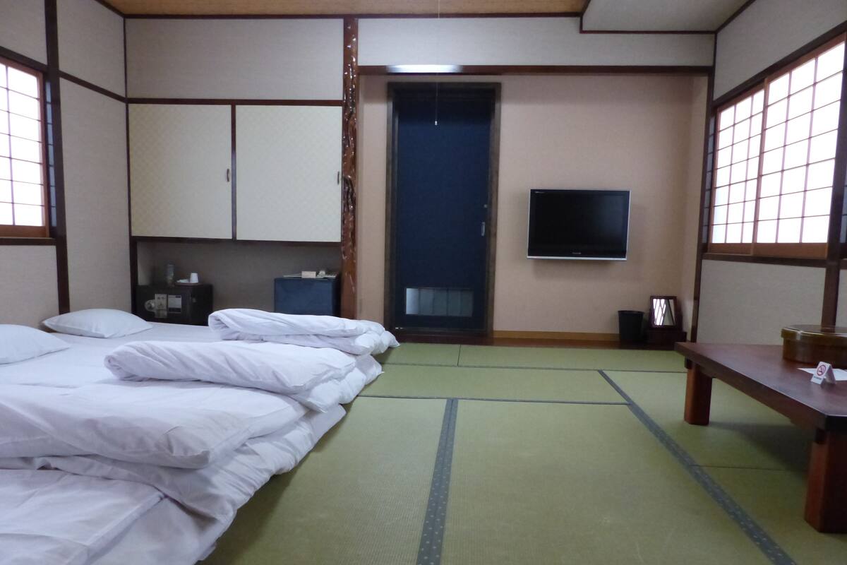 A traditional Japanese-style room with tatami mats, a bed with white bedding, a television on the wall, and a small table with a sign.