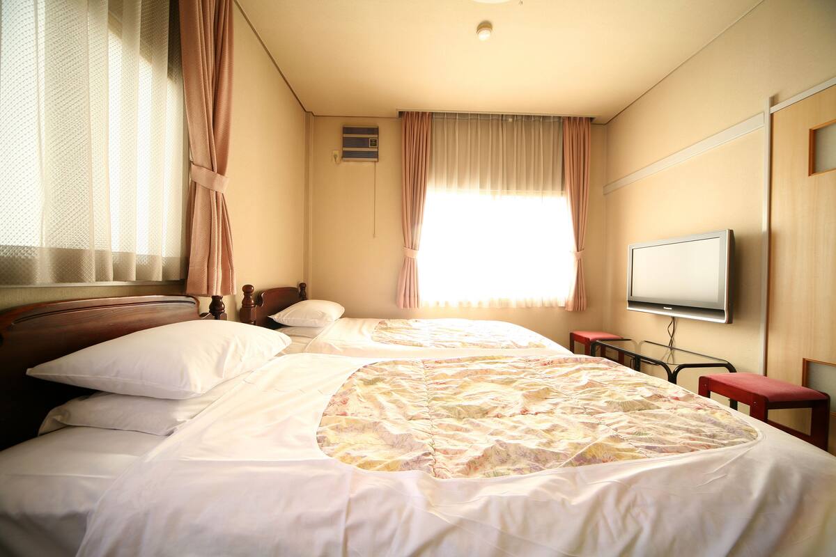 A hotel room with a bed, a television, a window with curtains, and a wooden headboard.