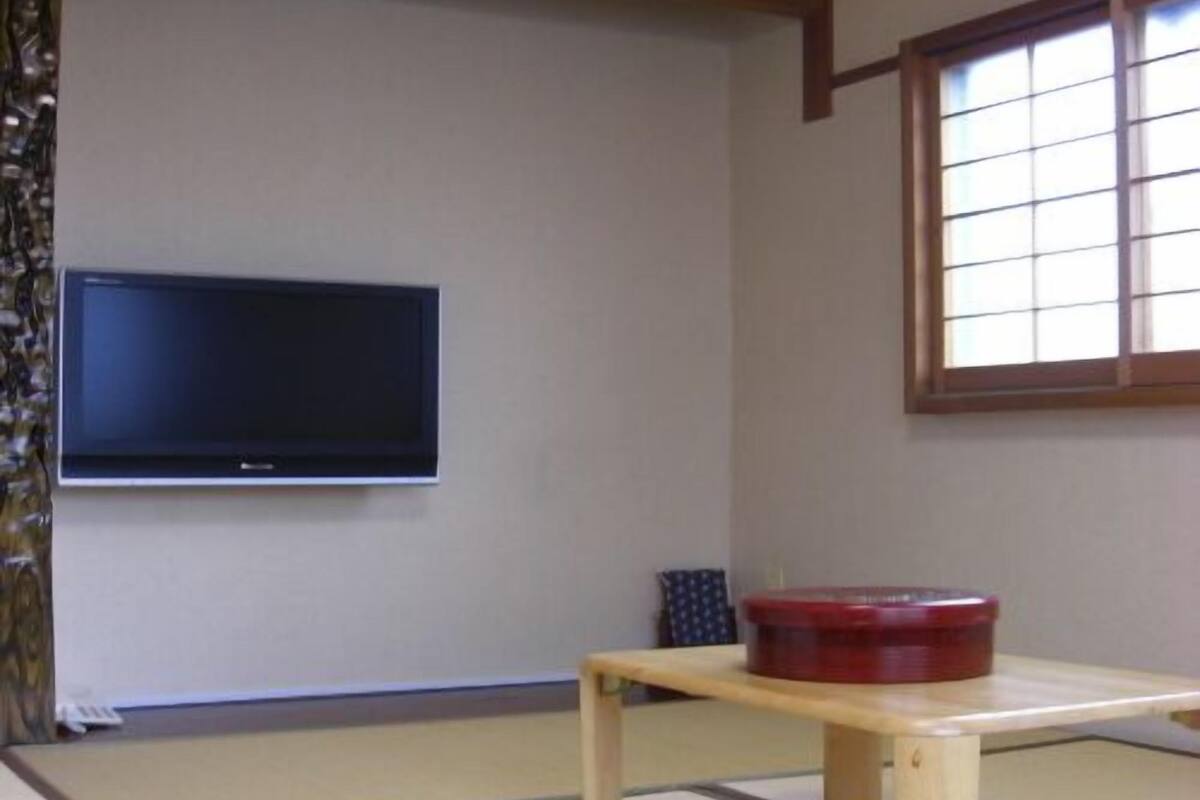 A tatami mat room with a television mounted on the wall, a low wooden table, and a window with a grid pattern.