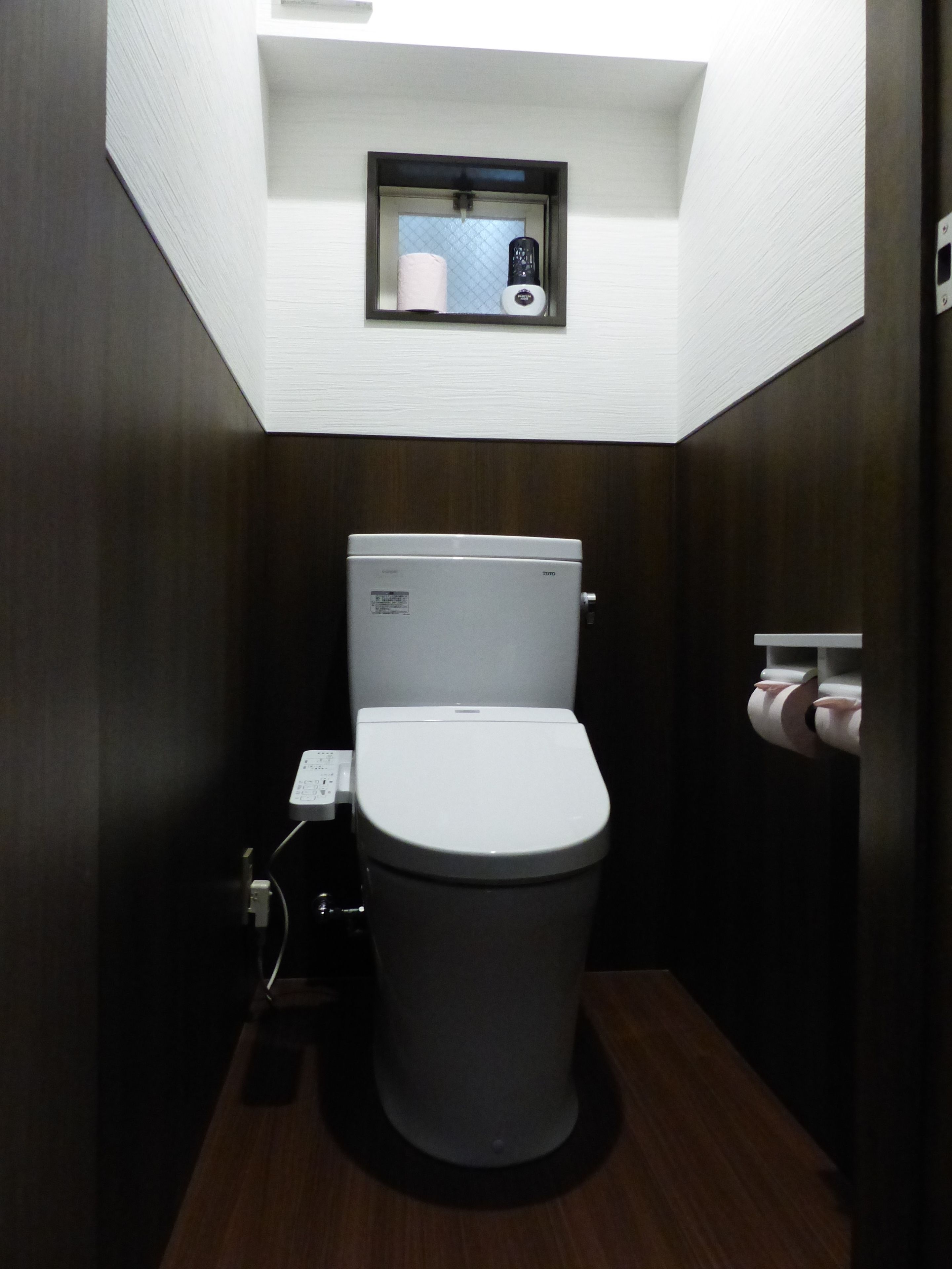 Casa de banho compacta e moderna, com sanita, uma pequena janela sobre a sanita e um rolo de papel higiénico fixado na parede.