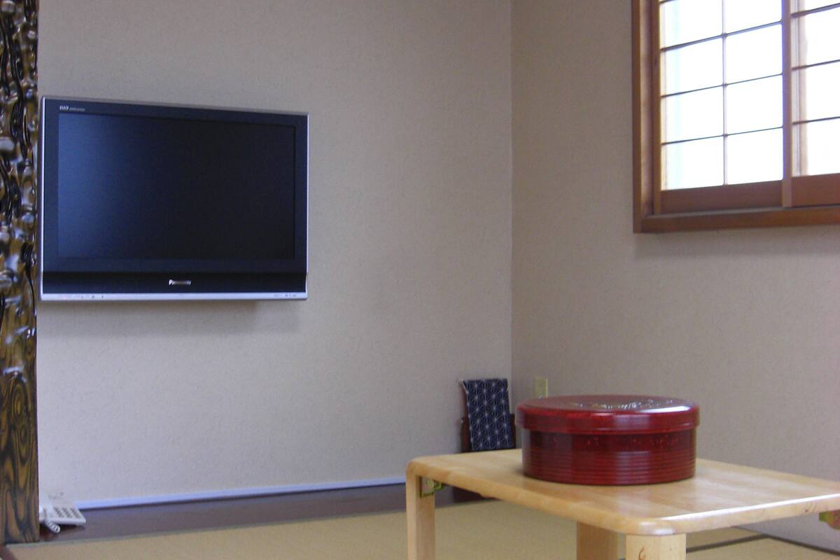 A traditional Japanese room with tatami flooring, a low wooden table, a television mounted on the wall, and a window with a grid pattern.